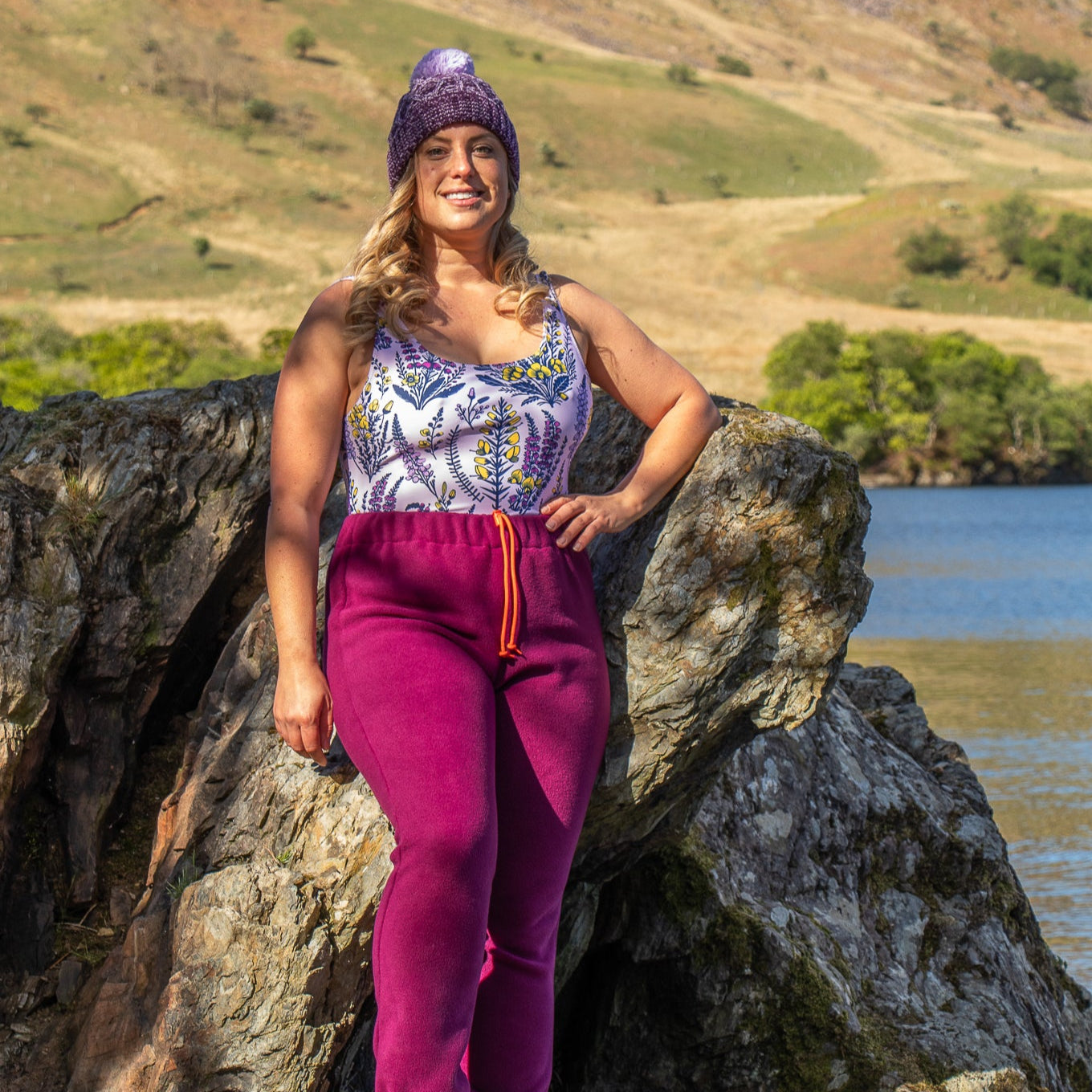 Woman in purple beanie and plum pants standing on a rock with a scenic background