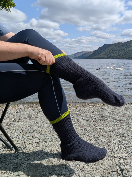 A person sitting outdoors by a lake wearing dark navy ribbed wool socks with bright green trim, paired with navy leggings, with mountains and water in the background.