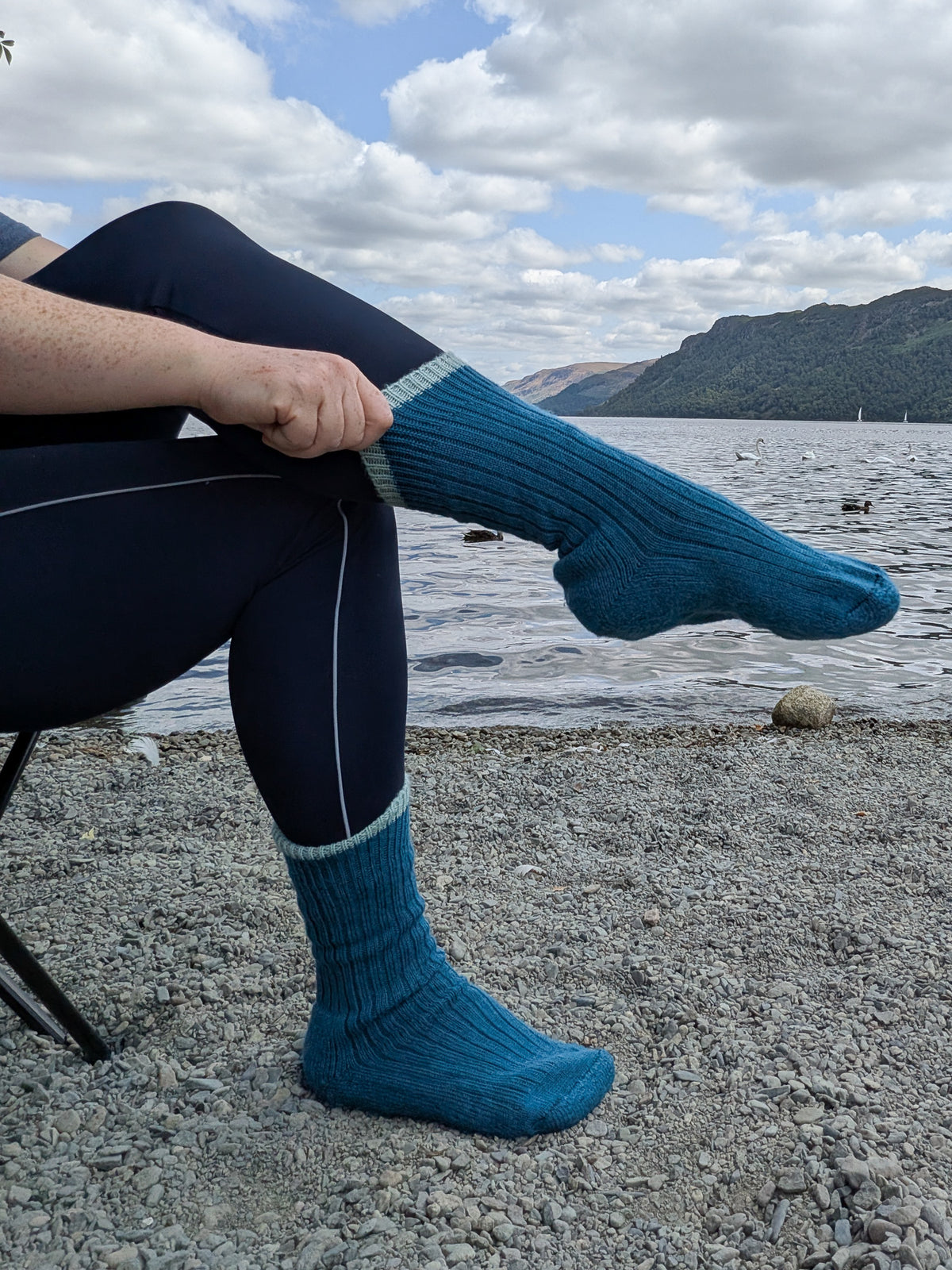 A person sitting outdoors by a lake wearing dark navy leggings and bright teal-blue wool knit socks, with mountains and cloudy sky in the background.