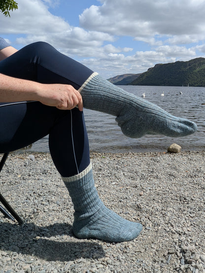A person sitting outdoors by a lake wearing dark navy leggings and light blue-gray ribbed knit wool socks, with mountains and cloudy sky in the background.